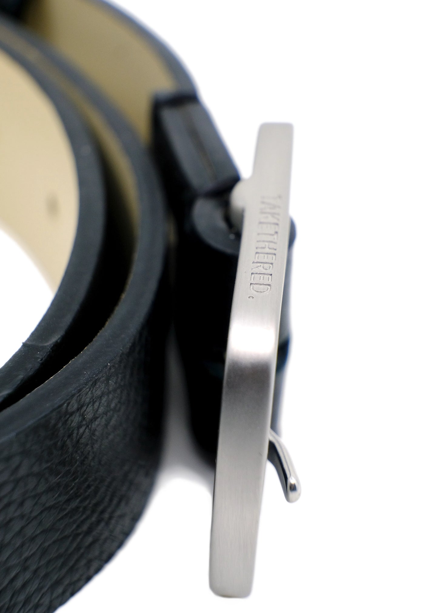Close-up of a weathered silver buckle engraved with "TAKETHERED." on a black pebble-grain leather Grunge Belt.
