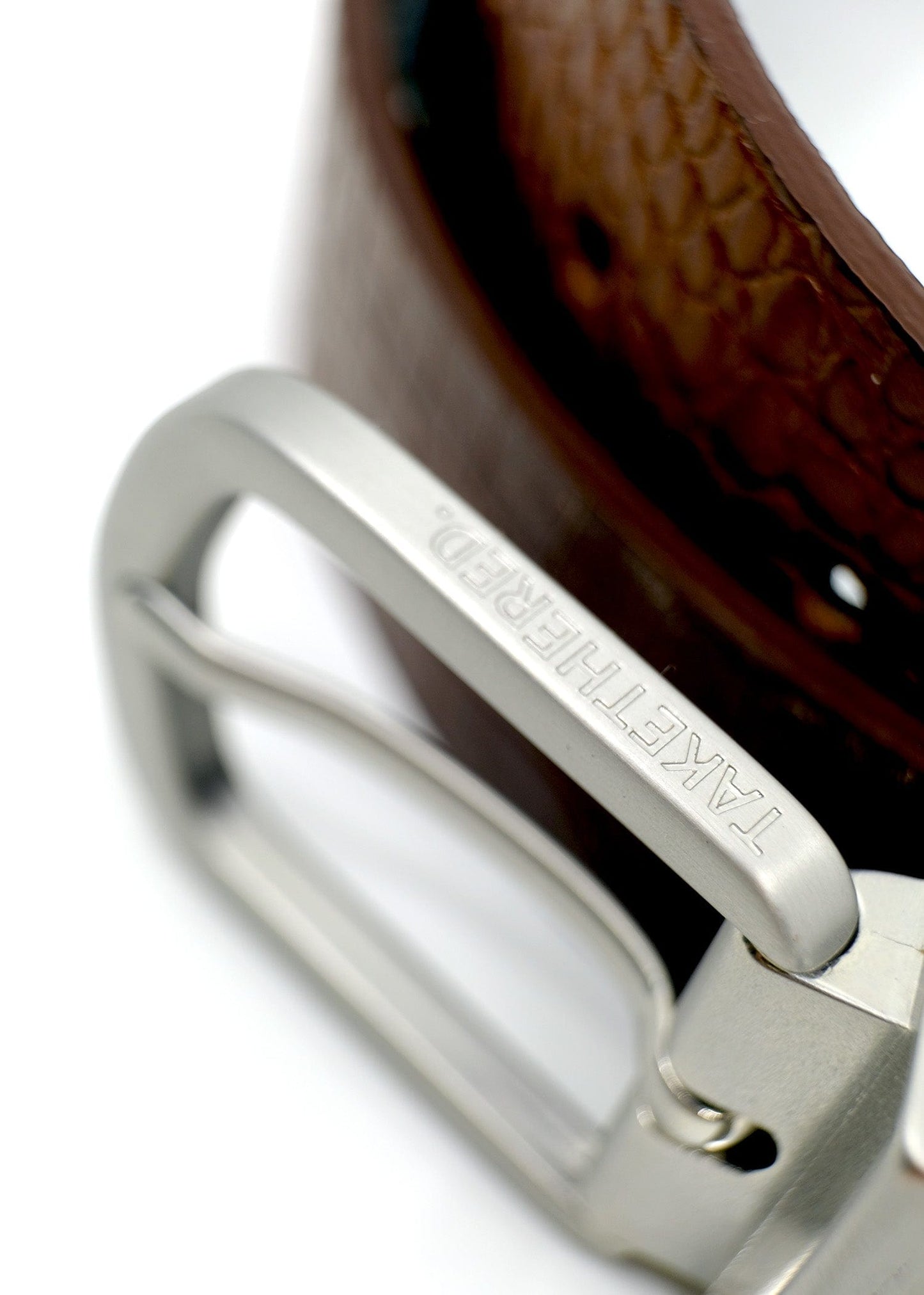 Extreme close-up of a reversible silver buckle with "TAKETHERED." engraved on the edge, featuring Smokey Cognac crocodile leather.