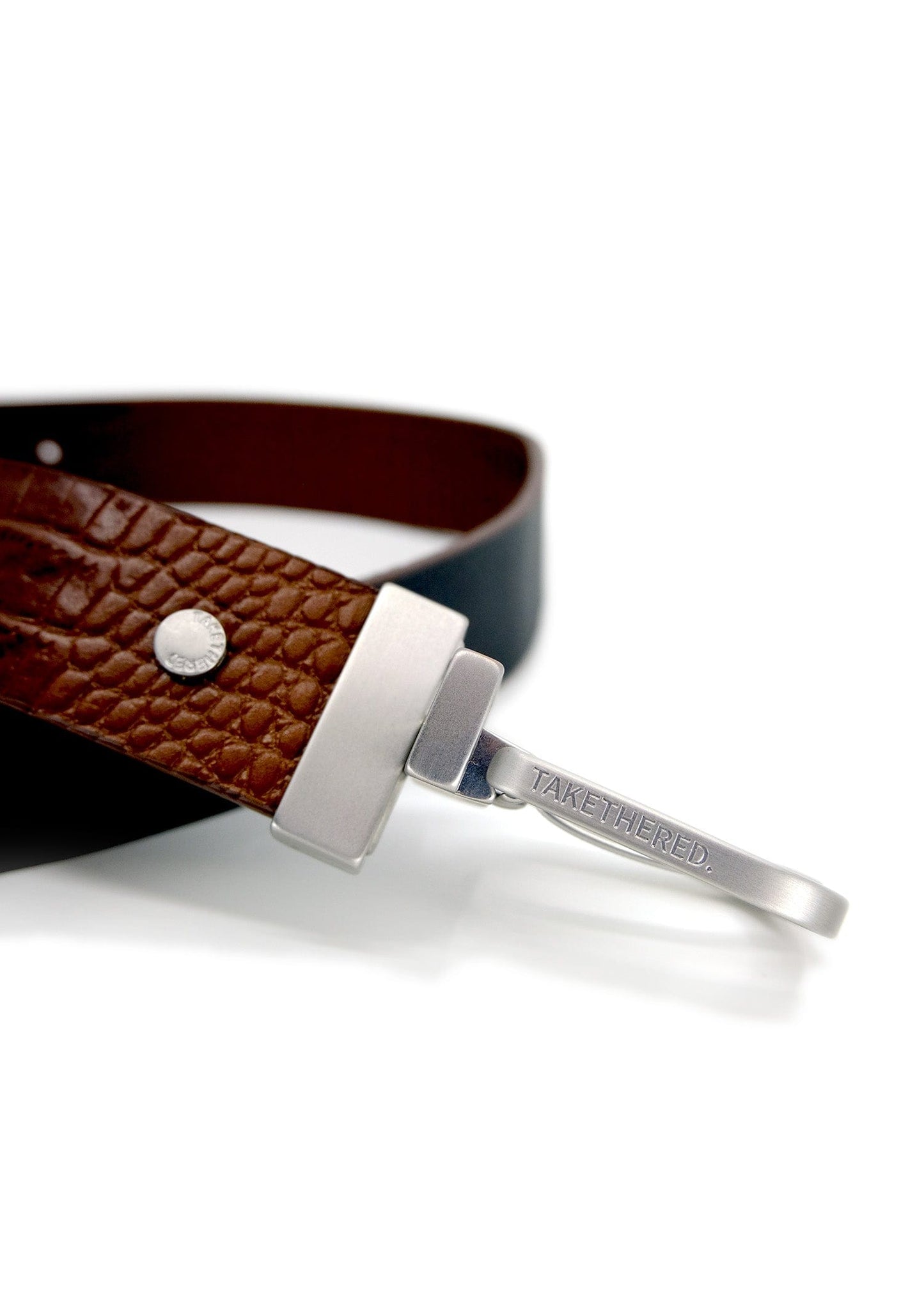 Close-up of Two-Face Belt with a reversible silver buckle engraved "TAKETHERED." and Smokey Cognac crocodile leather.