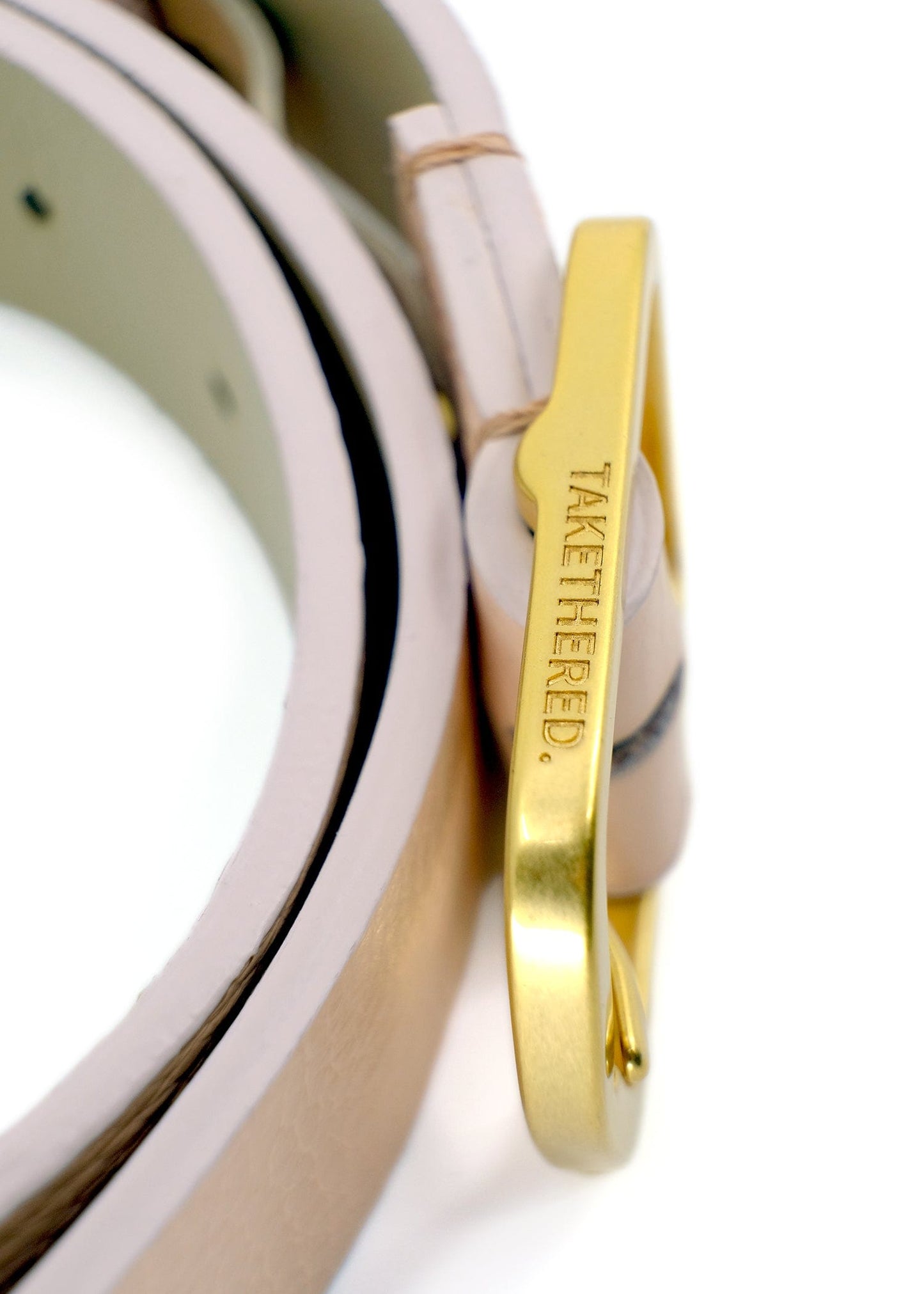 Side view close-up of a gold buckle engraved with "TAKETHERED." on a Nude Pink pebble-grain leather belt.