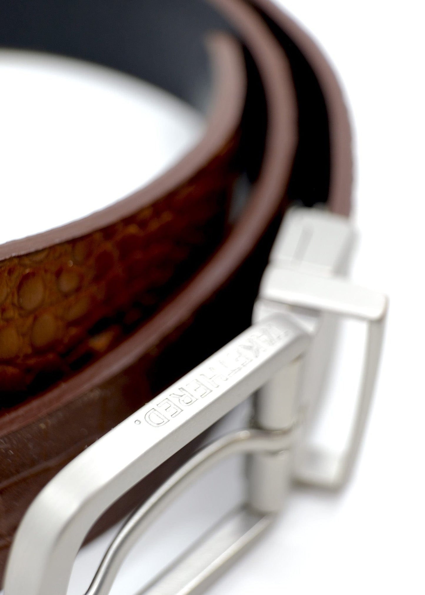 Extreme close-up of a reversible silver buckle with "TAKETHERED." engraved on the edge, featuring a coiled belt.