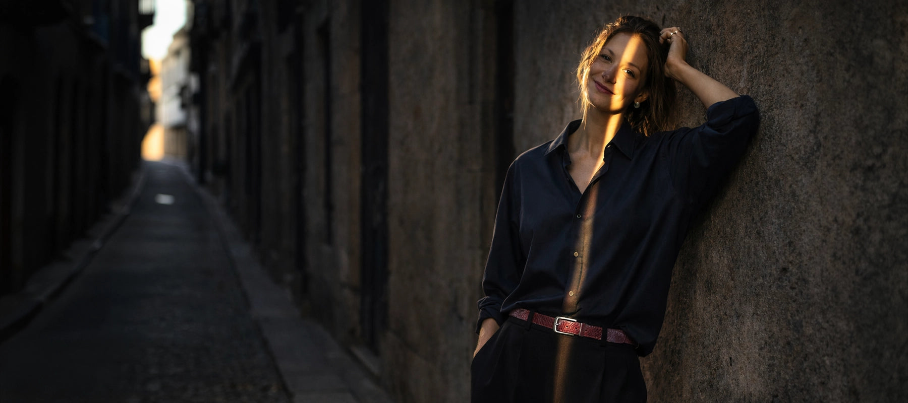 Woman leaning at a wall wearing  Serpent belt in Barbie Pink, black trousers and a dark blue shirt