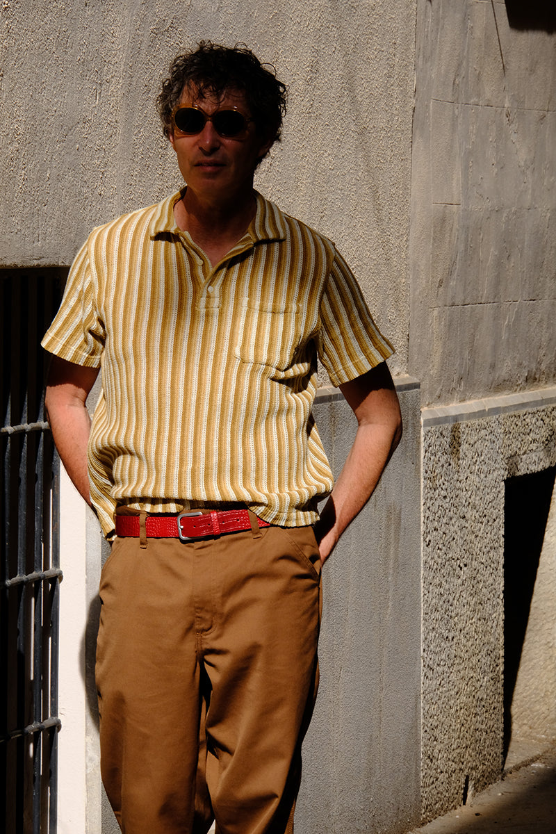 A man wearing a blood red Mississippi leather belt wearing a striped shirt, brown trousers, and walking in the sunshine.