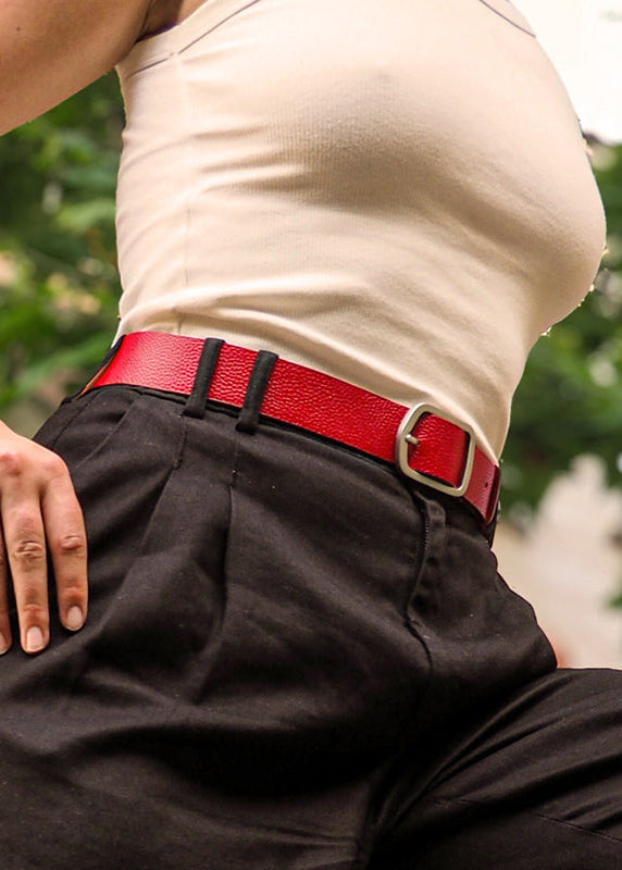 A knee shot of a womans waist wearin a blood red grunge belt in leather with a silver buckle.